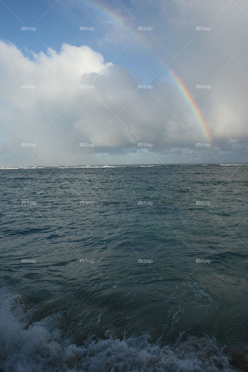 Rainbow, White Clouds & Surf. A rainbow rises over the ocean as waves hit the shore.