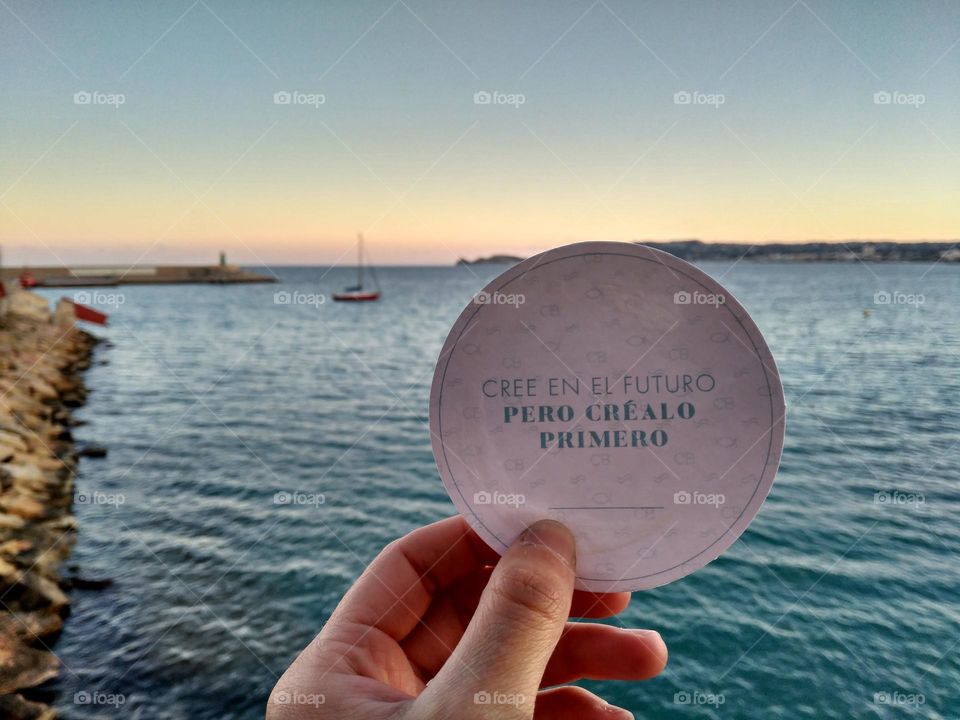 open blue sea with some boats and a "believe in the future but create it first" sign holding by a hand.