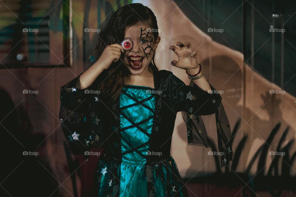 Portrait of a beautiful caucasian brunette girl in a witch dress holding a jelly candy in the shape of a human eye on her left eye smiling broadly and frightening with her second hand against the background of a painted wall at night on a school holi