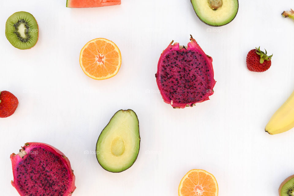 Flat lay of halved fruits scattered around a white tabletop including avocado, oranges, dragon fruit and watermelon