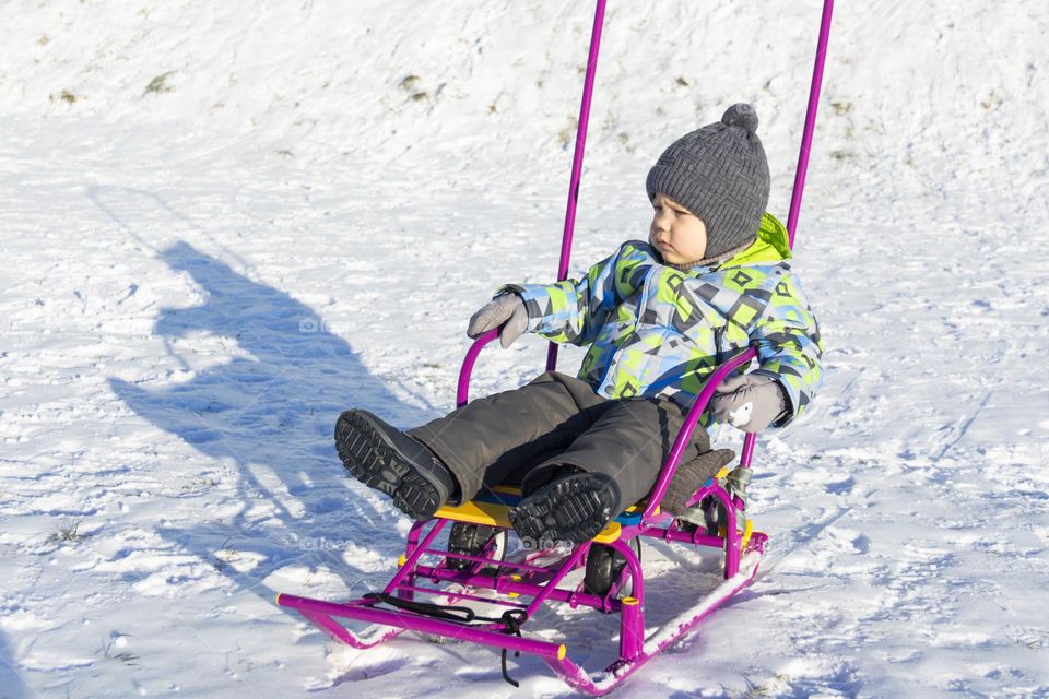 A child with a serious expression on his face in winter clothes jackets, pants, hat and boots in winter on white snow on the street and in the park in nature sledding and playing winter fun.