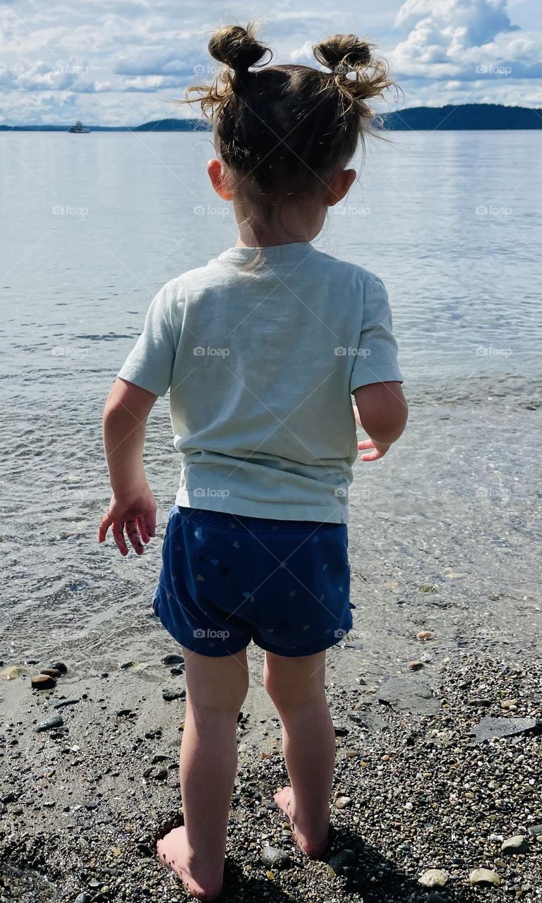 A toddler girl looks out towards the blue waters of Puget Sound from a sandy beach in Steilacoom, Washington 