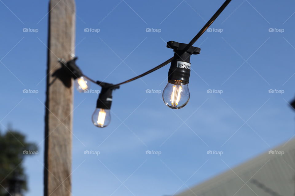 String of Glass electrical lightbulbs globes strung on wood post and illuminated in daytime against backdrop of blue sky