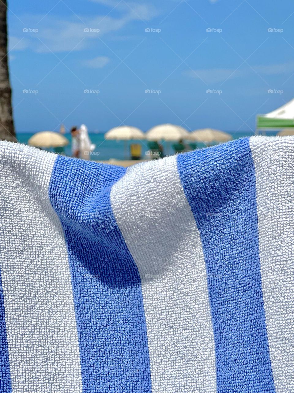 Blue and white striped hotel-issue beach towel strung up with a line of white beach umbrellas and visitors with the ocean behind them