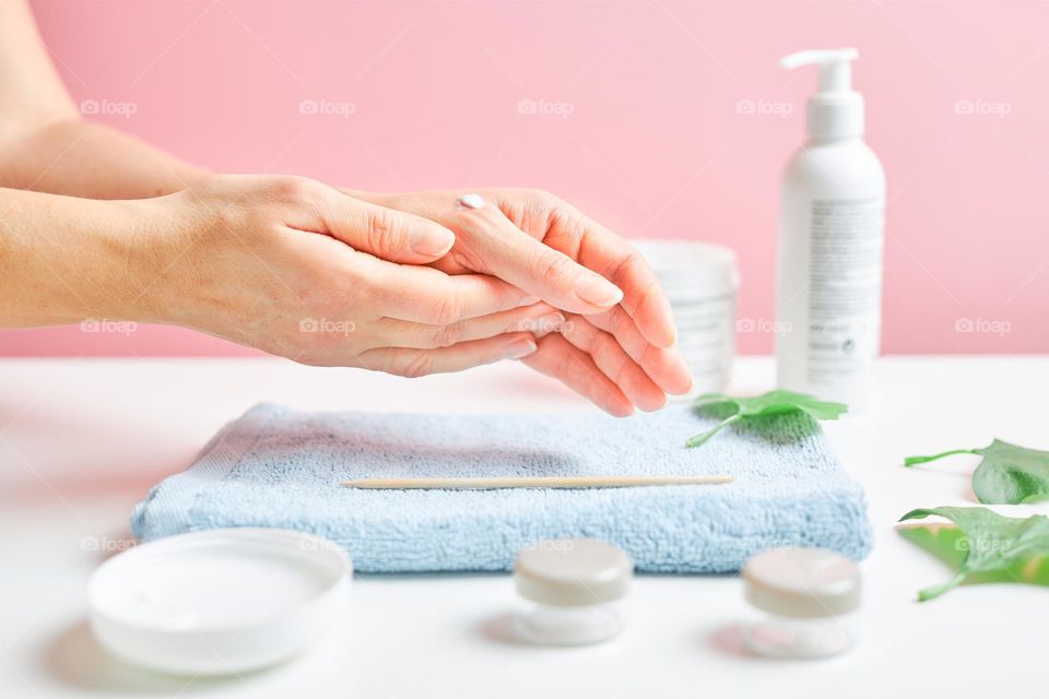Woman applying  cosmetic moisturizing hand cream. Cosmetic products, green leaves on white table. Spa, manicure, skin care concept. Flat lay, overhead view