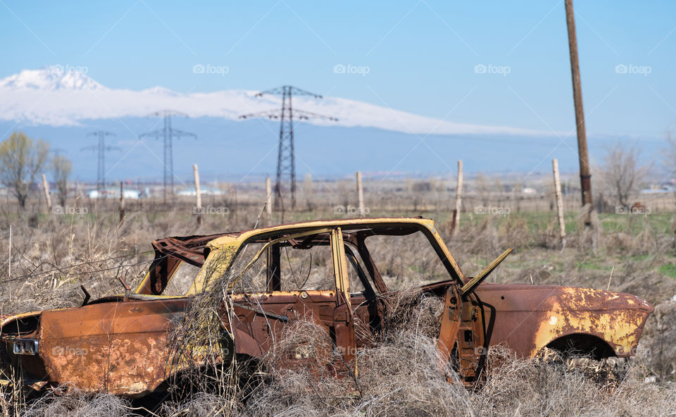 Abandoned and rusty wreckage of an yellow vintage Soviet Russian car in the middle of dry hay with scenic ice top mountains and clear blue sky on the background in rural Southern Armenia in Ararat province on 4 April 2017.