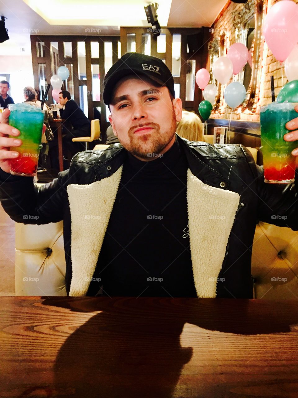 Gay pride photo of a male drinking two rainbow cocktails with pink and blue balloons 