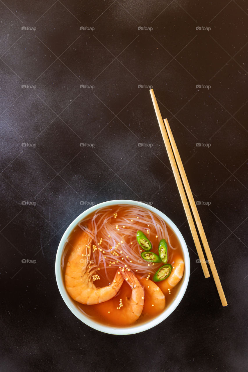 Top view of plate with asian soup made with shrimps, rice noodles, garlic and hot green pepper on black background with copy space.