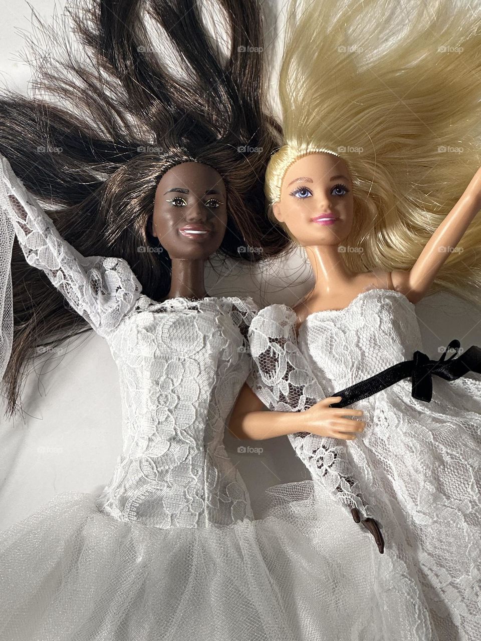 Two Barbies, one blonde and one brunette, are dressed in wedding white, celebrating as brides and waving to well wishers