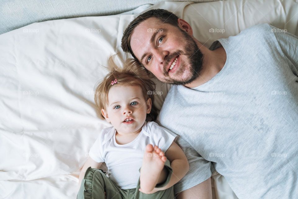 Young happy family with father and daughter baby girl on bed in cozy home, view from top