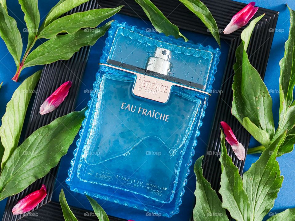 One glass blue transparent bottle of eau de parfum for men of the Versace brand lies in a black wooden frame with green leaves and bright pink peony petals on a blue background, flat lay close-up.