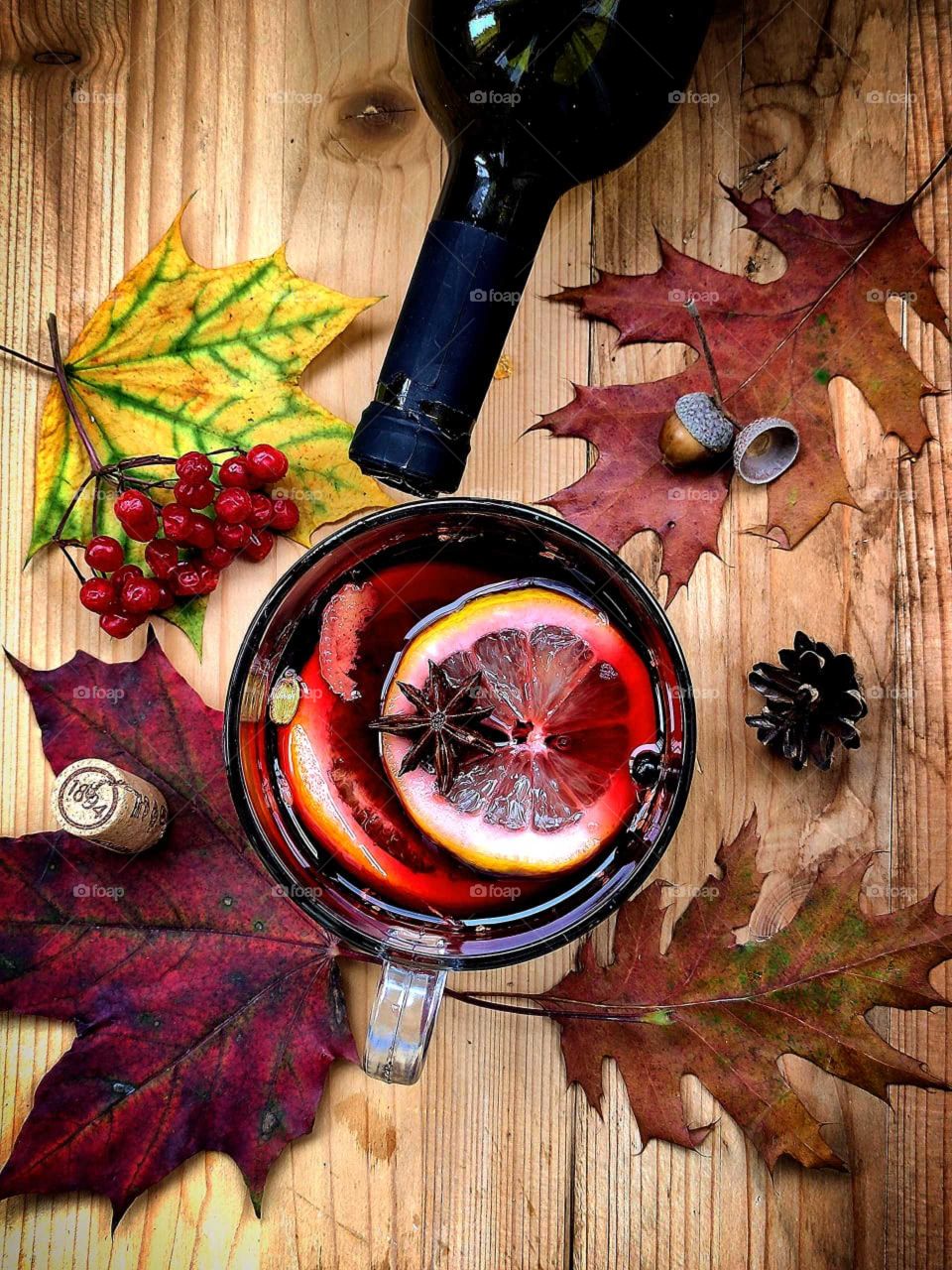 Composition.  A cup of mulled wine with lemon, orange and spices floating in it.  Next to the cup are: a red maple leaf with a wooden bottle cork, a yellow maple leaf with rowan berries, a dark bottle of wine