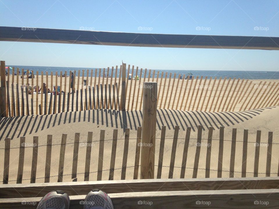 A view of the beach - and my sneakers- from a bench on the boardwalk in Point Pleasant Beach, NJ. The ocean is visible in the distance, and the fence on the dunes creates a pleasing pattern on the sand. People are on the beach enjoying the day. 