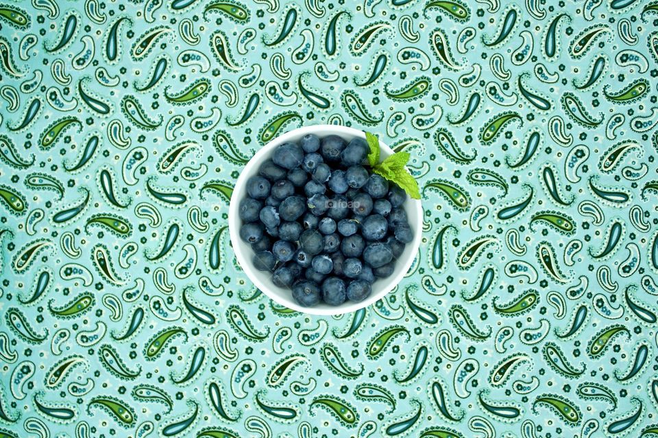 Blueberries and a spring of mint in a white bowl on teal and green paisley fabric