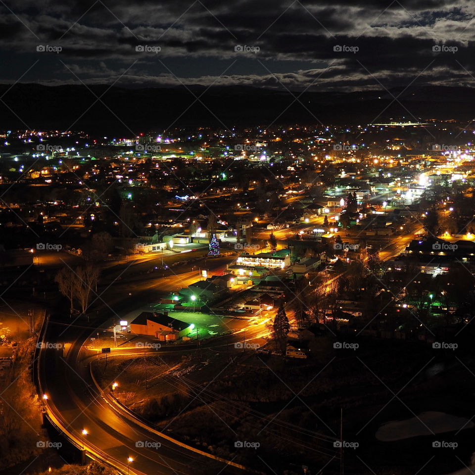 Beautiful glowing lights at night with holiday reds, greens, and golds looking down on the small town of Prineville in Central Oregon with its customary large Christmas Tree on the road coming into town and bright moonlight illuminating the clouds and sky above and silhouetting the surrounding hills. 