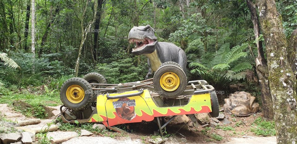 Dinosaur with wrecked vehicle in the woods.