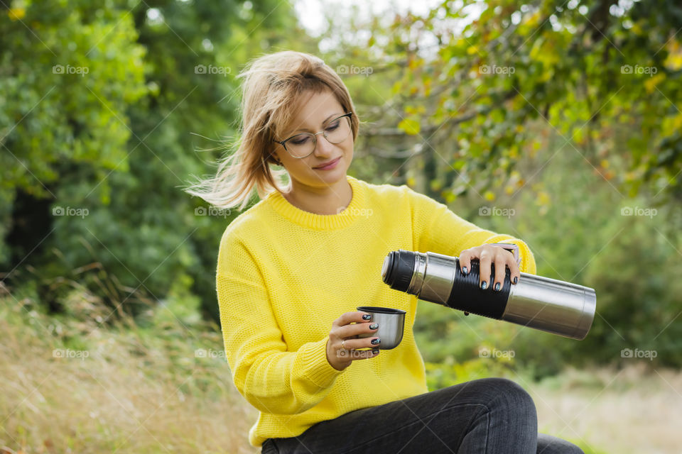 Beautiful woman in a cozy sweater sitting under a tree drinking hot drink tea outdoors in autumn at sunset light with bokeh fall cozy