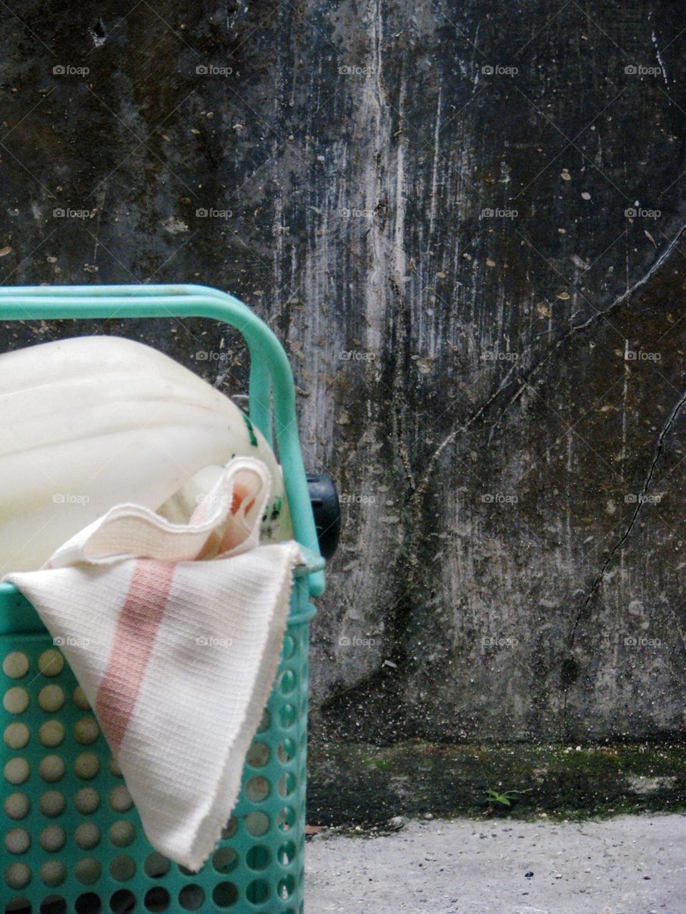 Close-up of a green plastic basket containing a cloth and a white helmet, placed in front of a dull wall and concrete floor