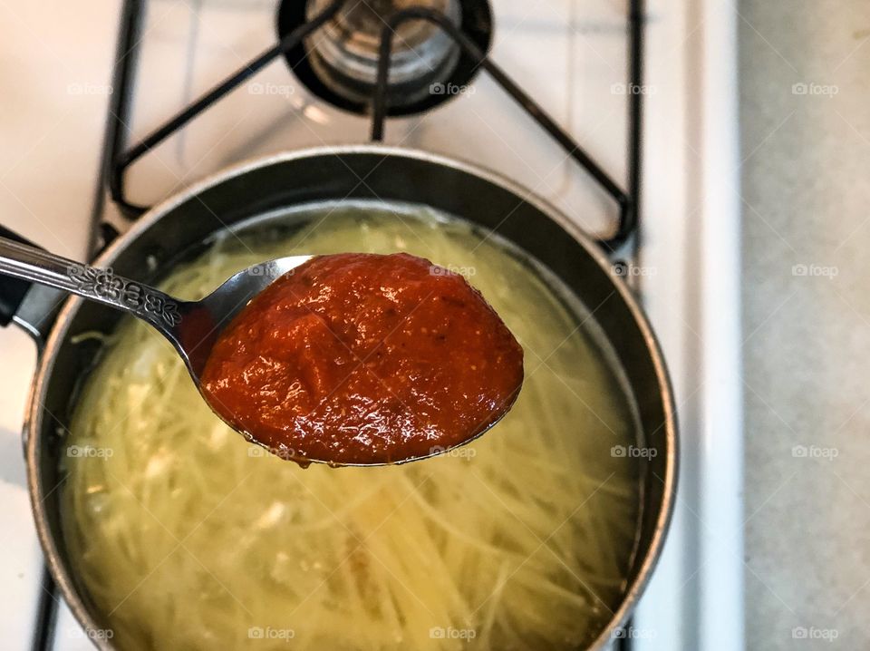 Fresh red spaghetti sauce in a spoon held directly above spaghetti noodles cooking below on a stove for dinner