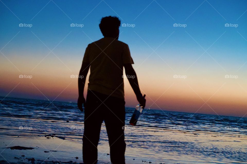 Standing on beach holding vodka bottle in the hand and looking at sunrise 