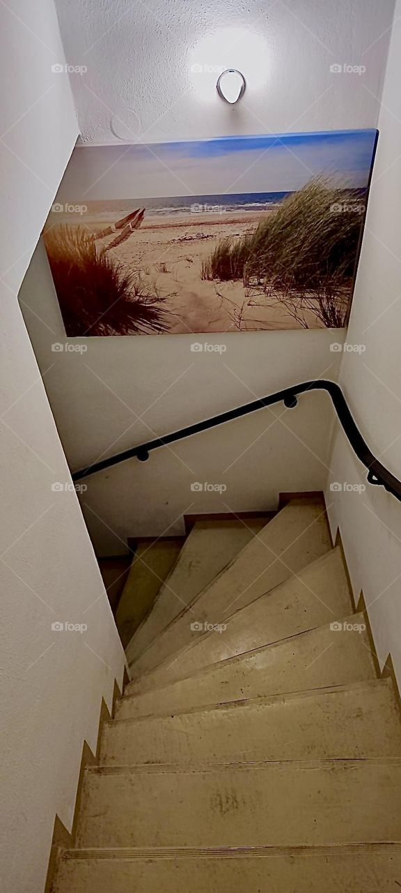 This is the staircase leading down to the subterranean recreation room in this one story bungalow in „Bavaria“, Germany. There is a railing and the walls are decorated with art. 2023. Hypnotic Productions