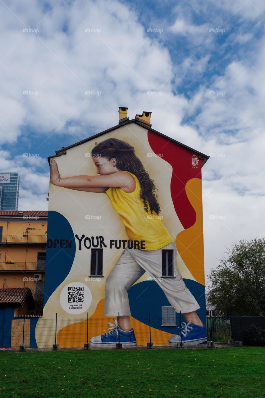 Milan, Italy colorful wall graffiti under blue sky. Vibrant street art mural by  artist Rosk depicting a young girl pushing a wall with the message Open Your Future.