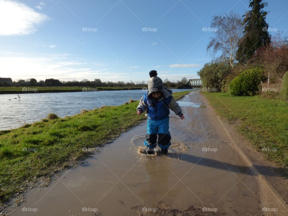 little boy dressed in warm blue clothes is jumping in the puddles by the river in early spring