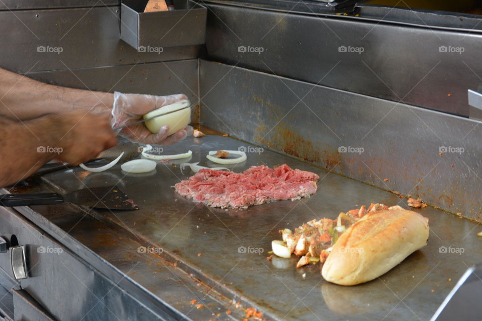 Hands are seen cutting onions on an open grill with meat, onions and bread on an outdoor street cart to sell food.