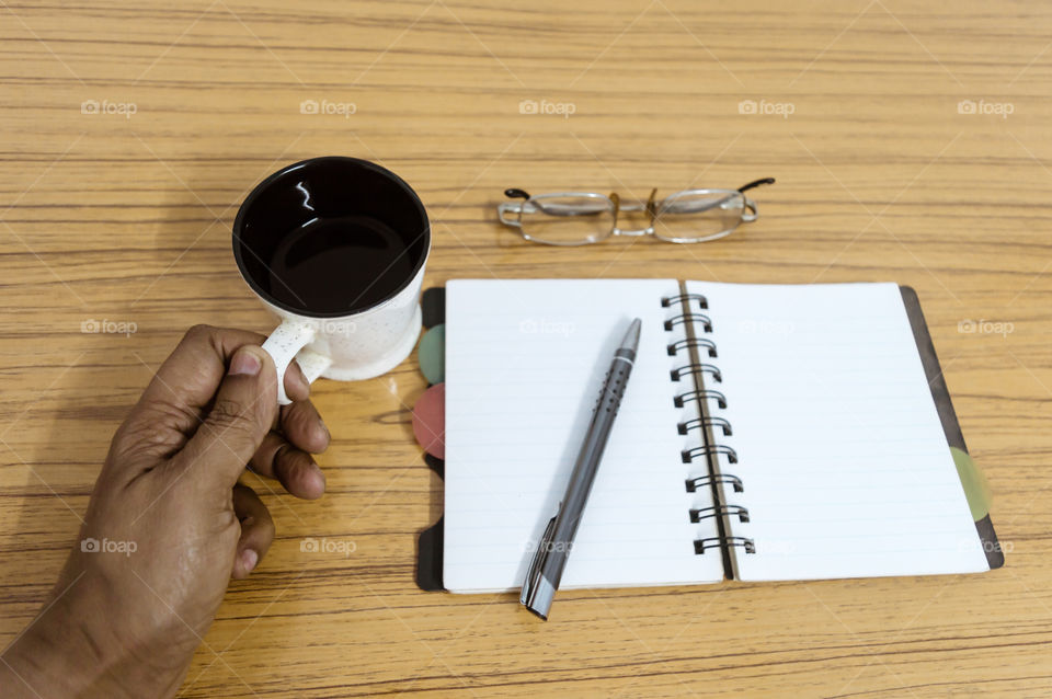 Businessman writing in his diary. Open notebook with blank pages next to cup of coffee and eyeglasses on wooden table. Business still life concept with office stuff on table. Top view with copy space.