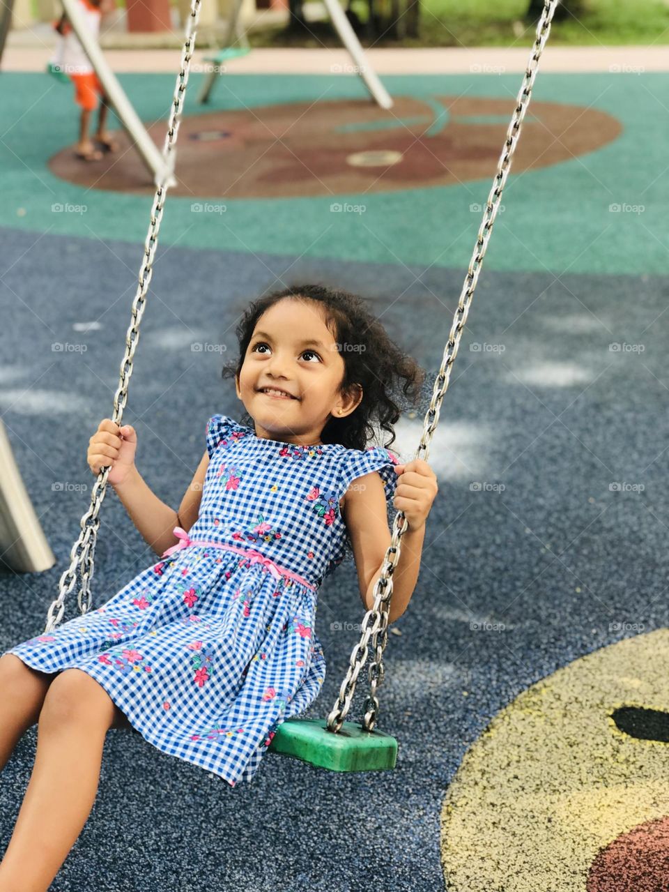 A four year old girl enjoying by swinging on swing and she looking upside and smiling and she wear blu colour frock.