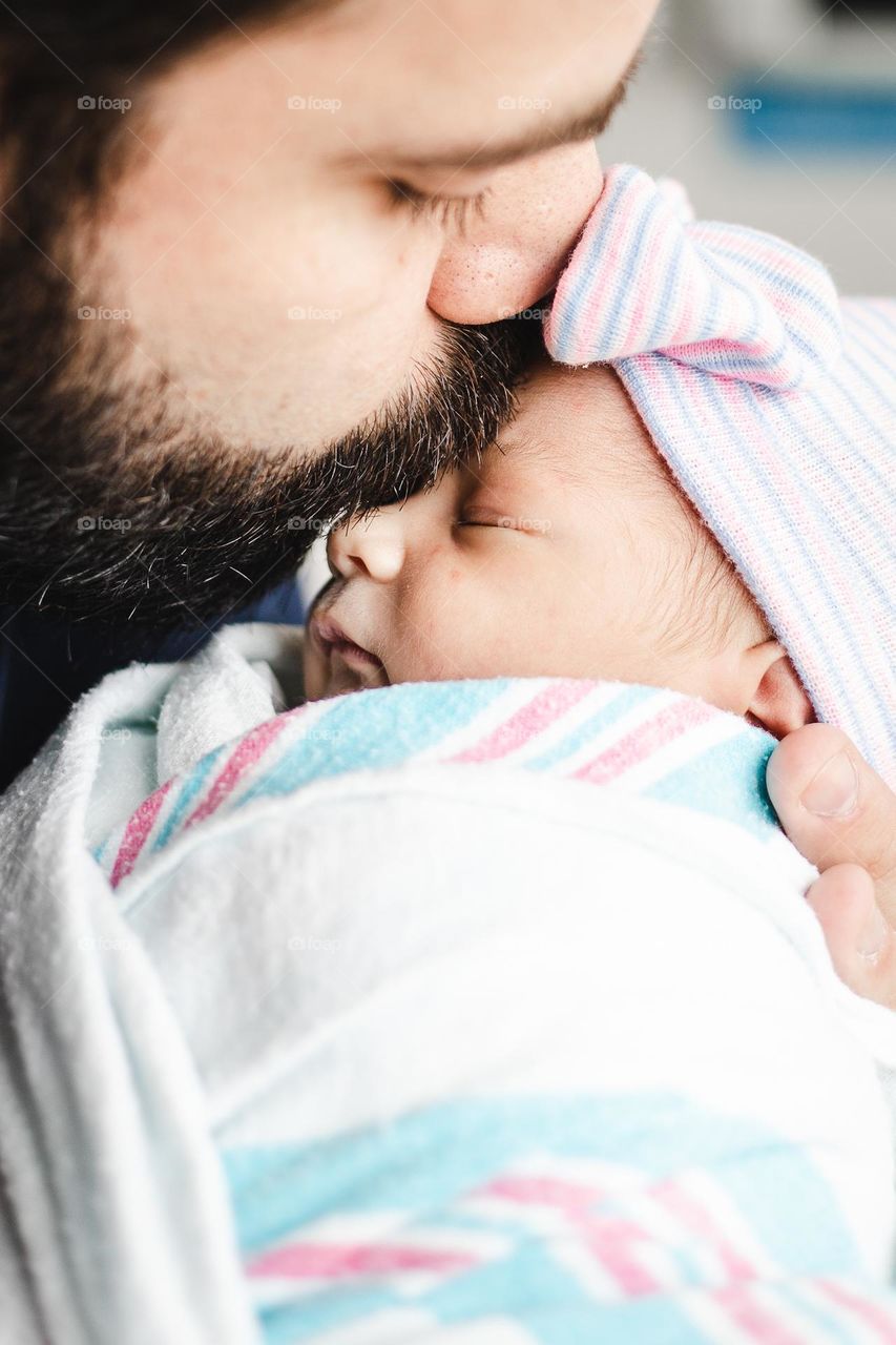 Close up of a new father kissing his newborn girl for the first time while she is wrapped in her hospital blanket and warm hat.