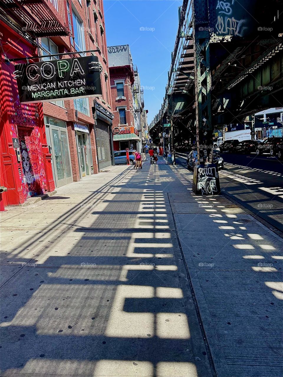 This is “Broadway” in “Bushwick”, Brooklyn. We are walking along the MTA “M” train overpass towards the “Myrtle Avenue” station and “Jefferson Street”. The overpass casts dramatic shadows onto the sidewalk. 2024. Hypnotic Productions