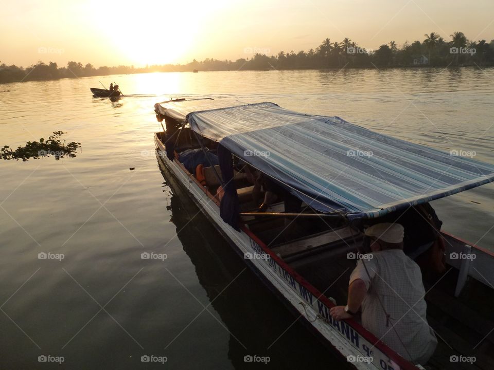 sitting on a covered boat going to floating market on the river early morning,the sun is radiating an orange color in the sky, reflecting its light into the river and trees.makes a wonderful sight