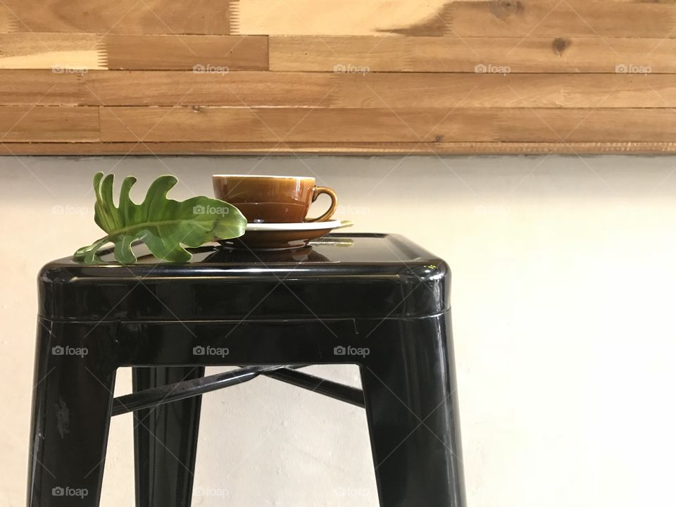 brown cup of hot coffee with bottom plate and green plant part on black metal bar stool with pieces of natural wood as coffee bar and white cement background