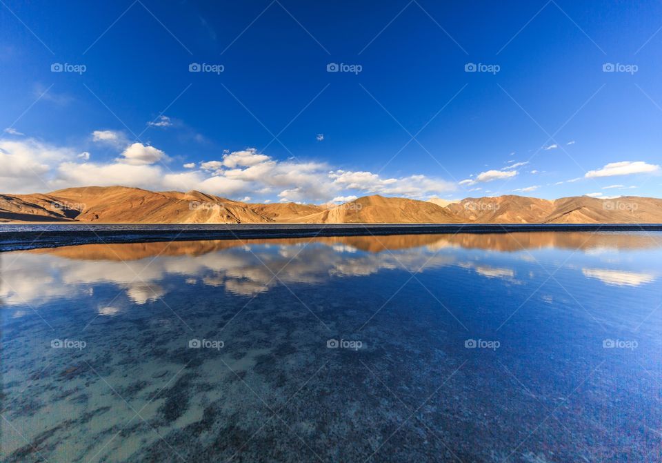 reflection of puffy white clouds and brown mountains on the frozen lake water