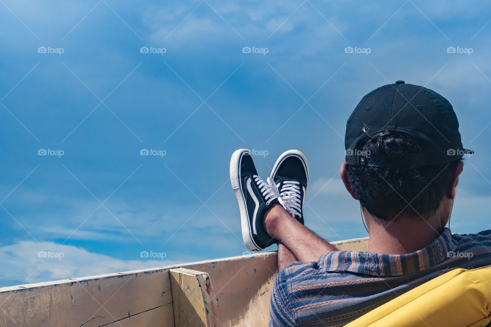 A rear view of a man sitting down on a front side of a boat, relaxing his feet, enjoying the fresh air
