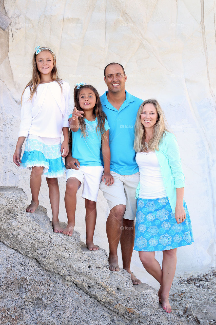 Young family of four, mom, dad, kids, daughters, children in blue and white casual clothes standing in a line on top of rocks outdoors at the beach