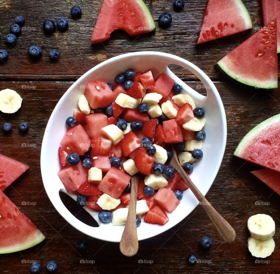 Fruit salad made with fresh watermelon, blueberries and banana in a white bowl with wooden serving spoons on a rustic wood table.