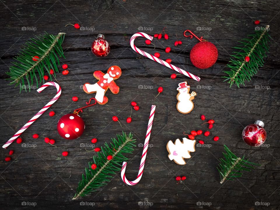 Flat lay of red and white Christmas decorations consisting of candy canes,gingerbread,red ribbon, red globes, evergreen branches and red berries on rustic wooden table 
