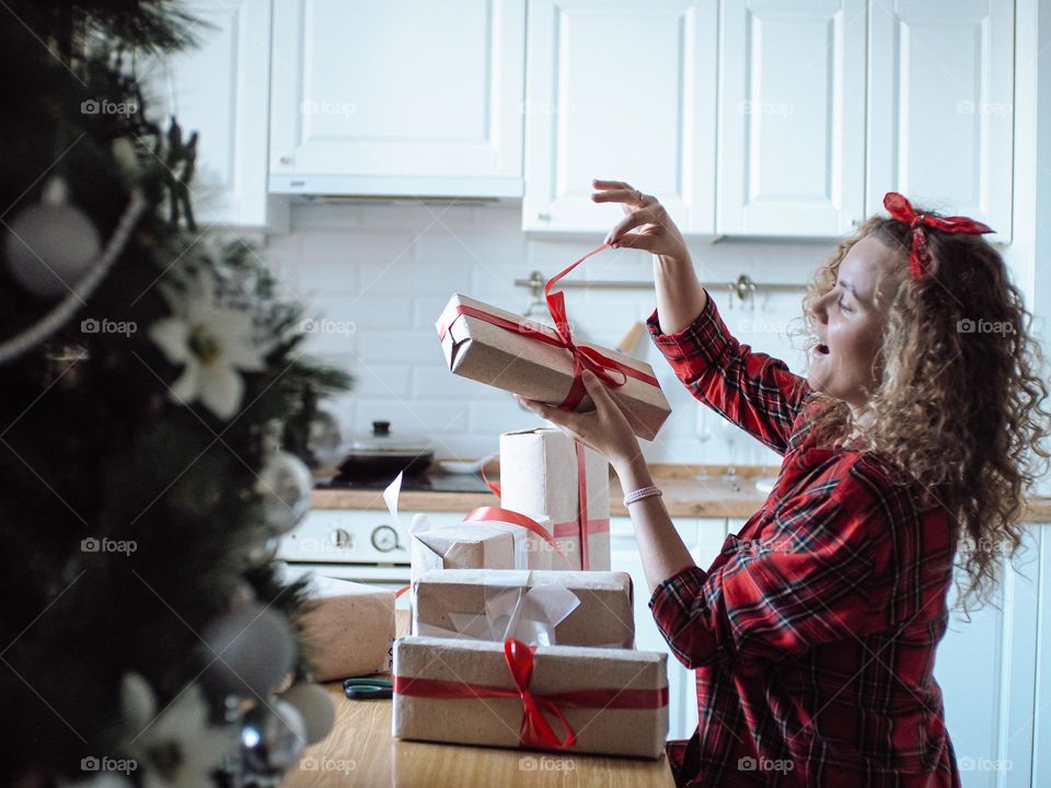 Young beautiful woman with curly hair in plaid shirt wrapping Christmas presents with red ribbons 