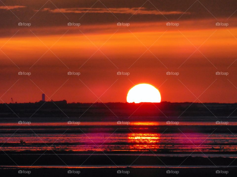 A reddish-golden sun, melting into the semi-horizon. Silhouettes mask the fire-coloured sky, with layers of shadows and sea. A glistening sunset reflection can be seen 