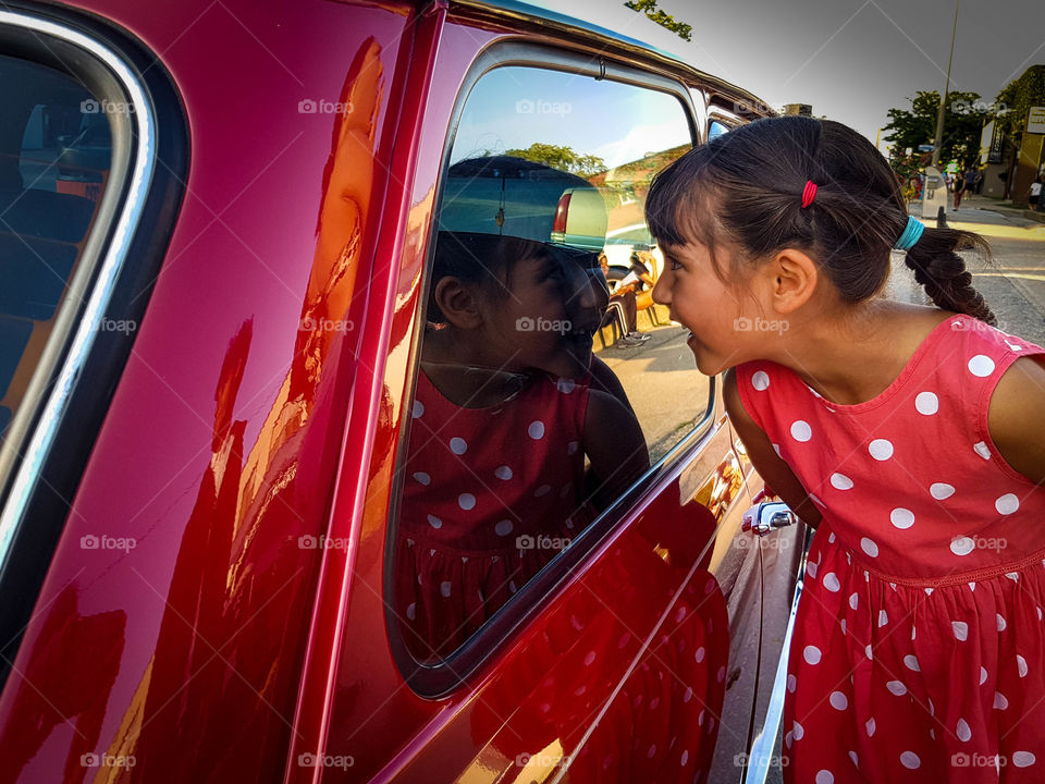 Cute little girl in a red dress is looking into a red car's passenger window