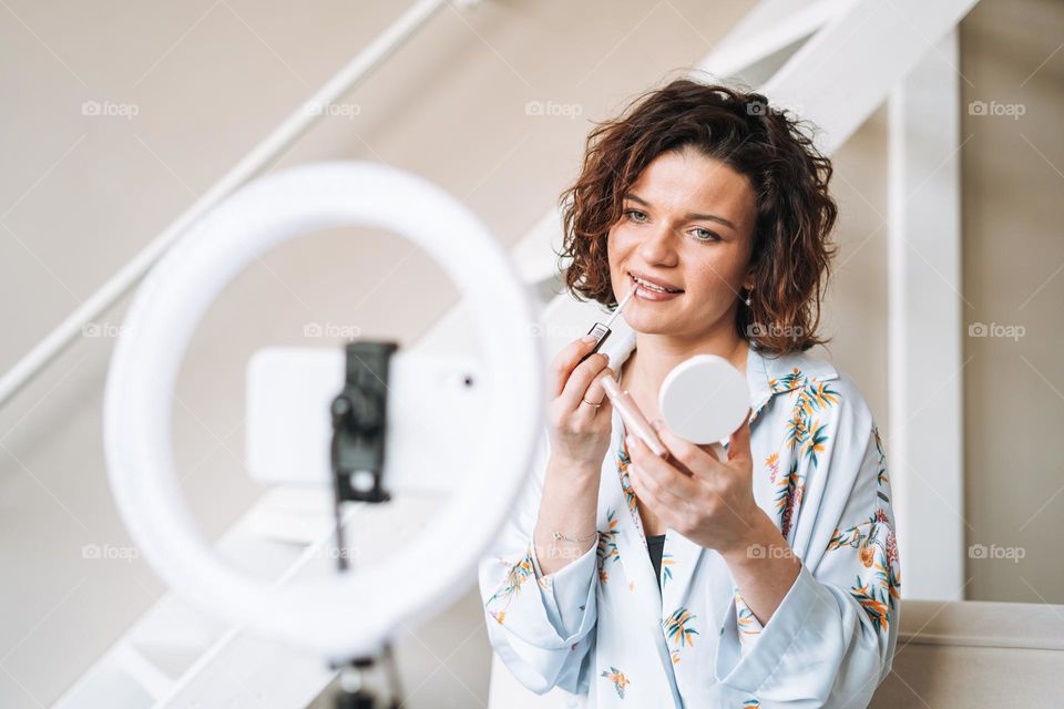 Young brunette woman blogger in home clothes doing makeup with mobile phone and ring lamp online in studio