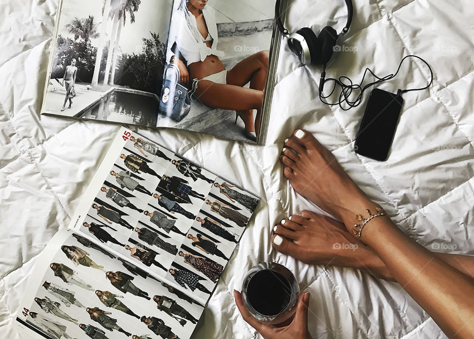 Young woman enjoying her weekend with a glass of wine, modern magazines and music through the headphones in cozy bed at home 