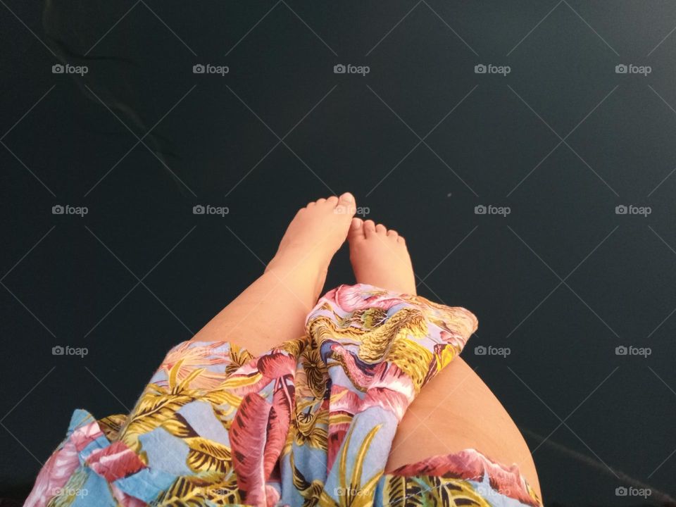 Female Adult with pretty Feet and Legs, dangling them over Water and pointing Toes towards the Ocean, her Colourful and multicoloured Flower patterned Dress, seen just covering her knees.