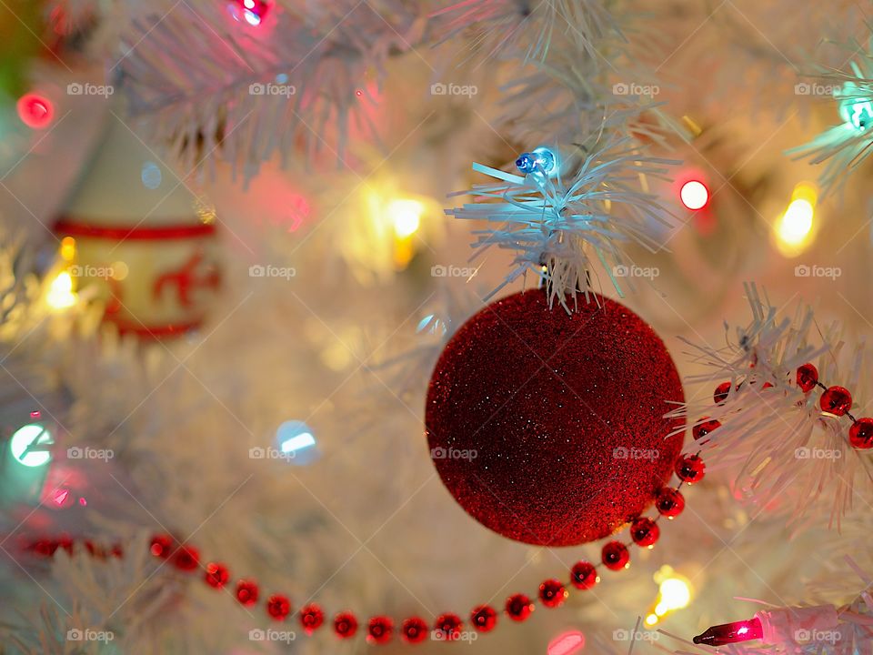 A glittery round red ornament hangs from a white Christmas tree with soft colored lights. 
