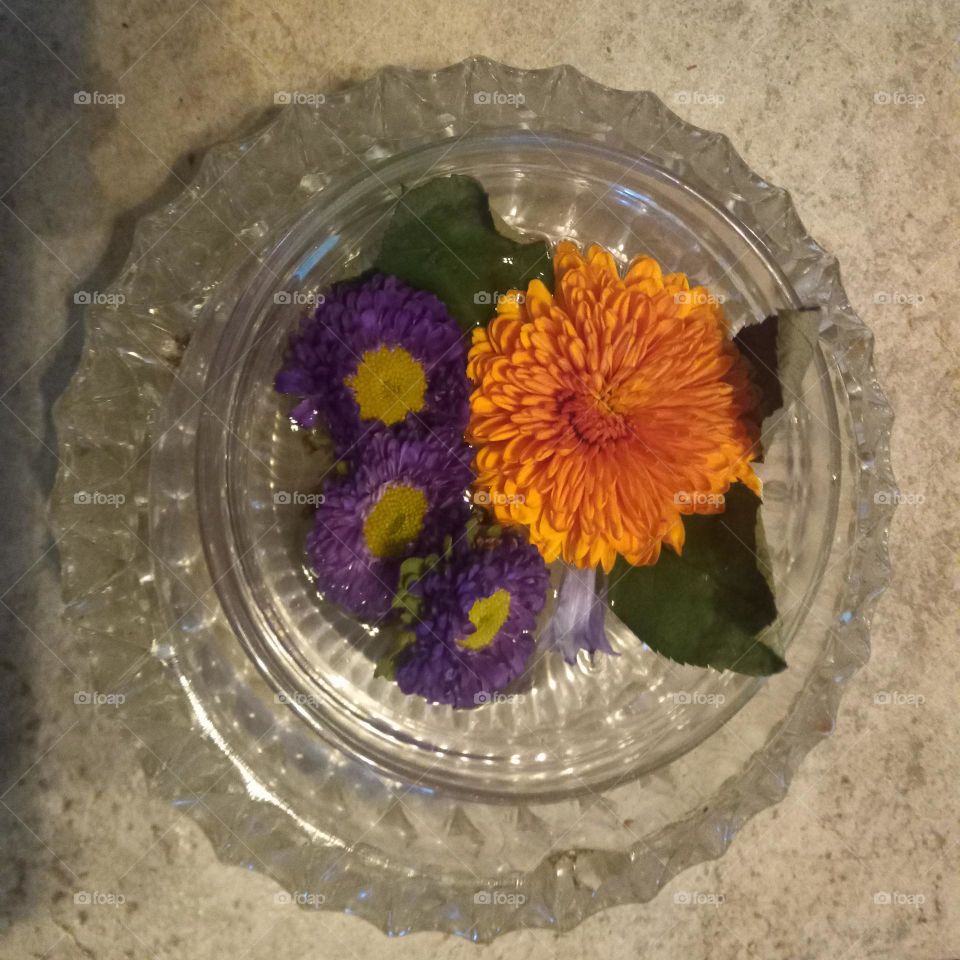 An ornate crystal dish setting on a brown and white swirled tile with a lovely orange and white flower  focus and small purple flowers and green accent leaves to compliment.