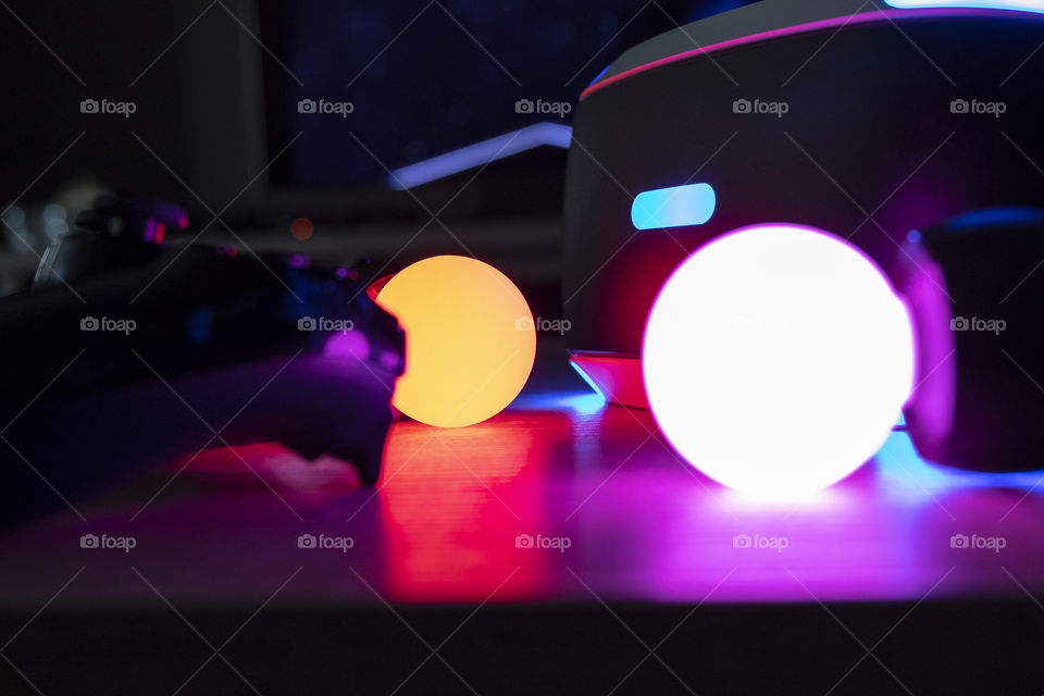 A portrait of turned on PlayStation move controllers, a PlayStation 4 controller and a virtual reality headset. it is a very colorful scene in a dark room.