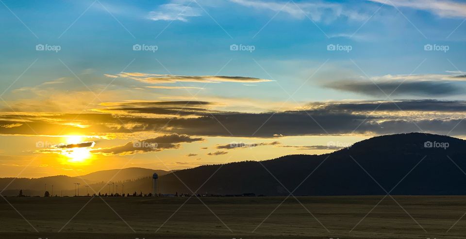 Bright yellow sun setting behind a sleepy and small farm town. Light and bright blue sky show the silhouette of the small town and the mountain range it is named for. 