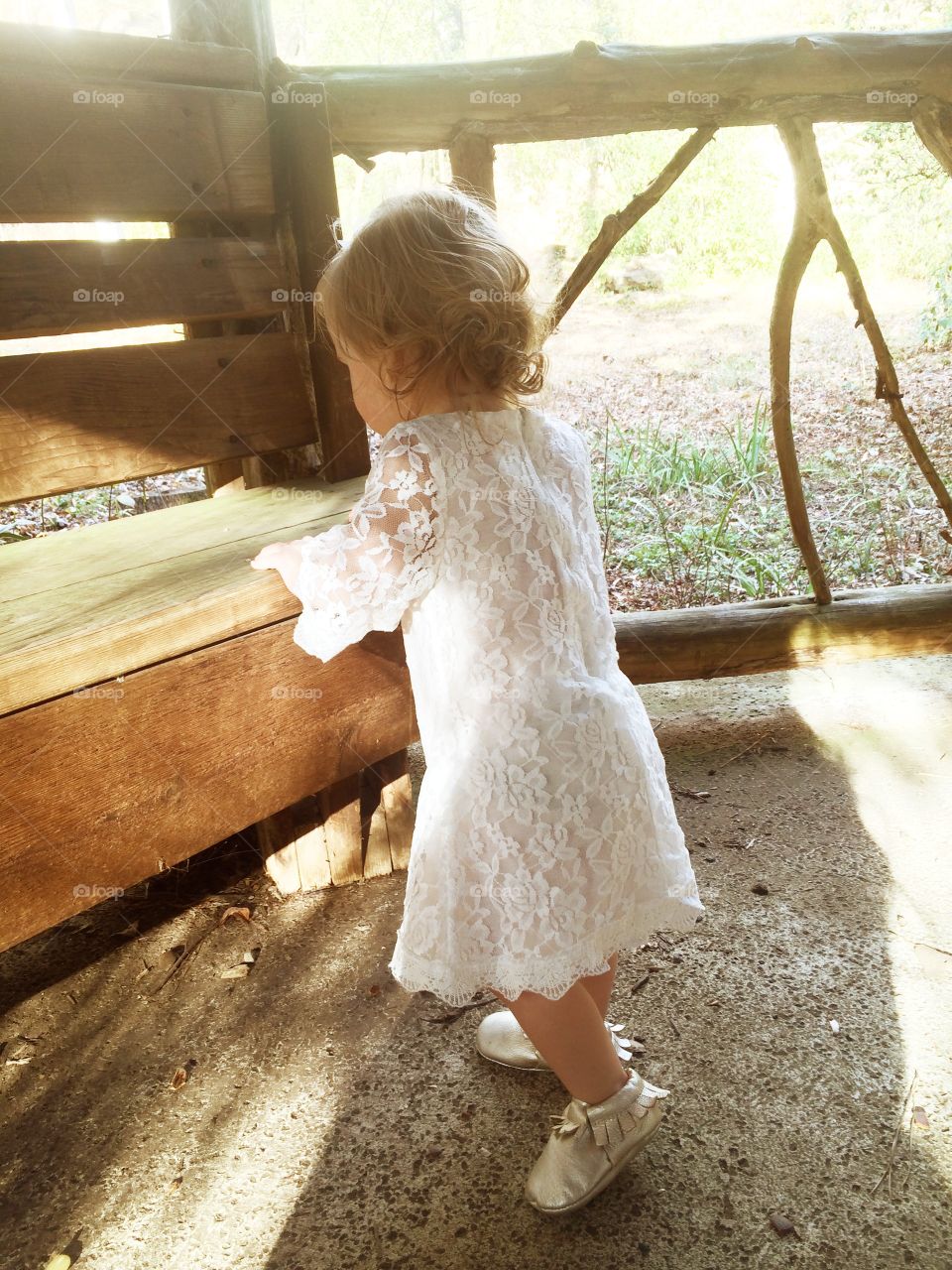 Toddler baby girl with curly blonde hair in white lace dress by wooden bench